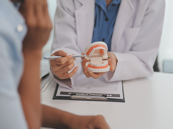 A dental professional examines a patient s teeth with a dental mirror while seated at a desk.